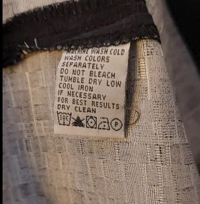 May include: Close-up of a clothing tag with care instructions. The tag is white with black text, including "MACHINE WASH COLD," "TUMBLE DRY LOW," and "DRY CLEAN." The fabric has a textured, woven pattern in shades of gray and white.