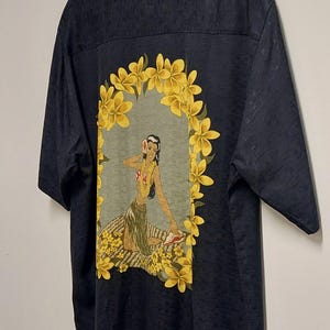 May include: Dark navy short-sleeve shirt with a Hawaiian-themed design. The back features a graphic of a woman surrounded by yellow flowers. The shirt has a subtle textured pattern.