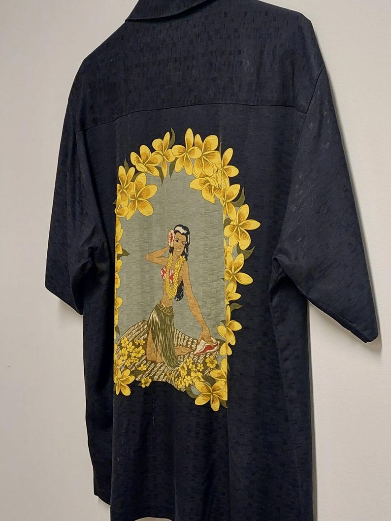 May include: Dark navy short-sleeve shirt with a Hawaiian-themed design. The back features a graphic of a woman surrounded by yellow flowers. The shirt has a subtle textured pattern.