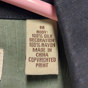 May include: Close-up of a tag from a silk shirt. The tag is beige with brown text that reads "M BODY: 100% SILK DECORATION: 100% RAYON MADE IN CHINA COPYRIGHTED PRINT."