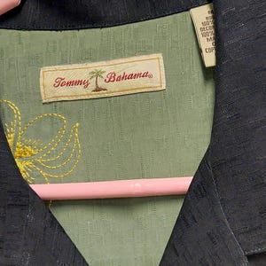 May include: Close-up of a Tommy Bahama shirt. The shirt is a dark navy blue with a green interior. The interior has a floral design with the Tommy Bahama logo in the center.