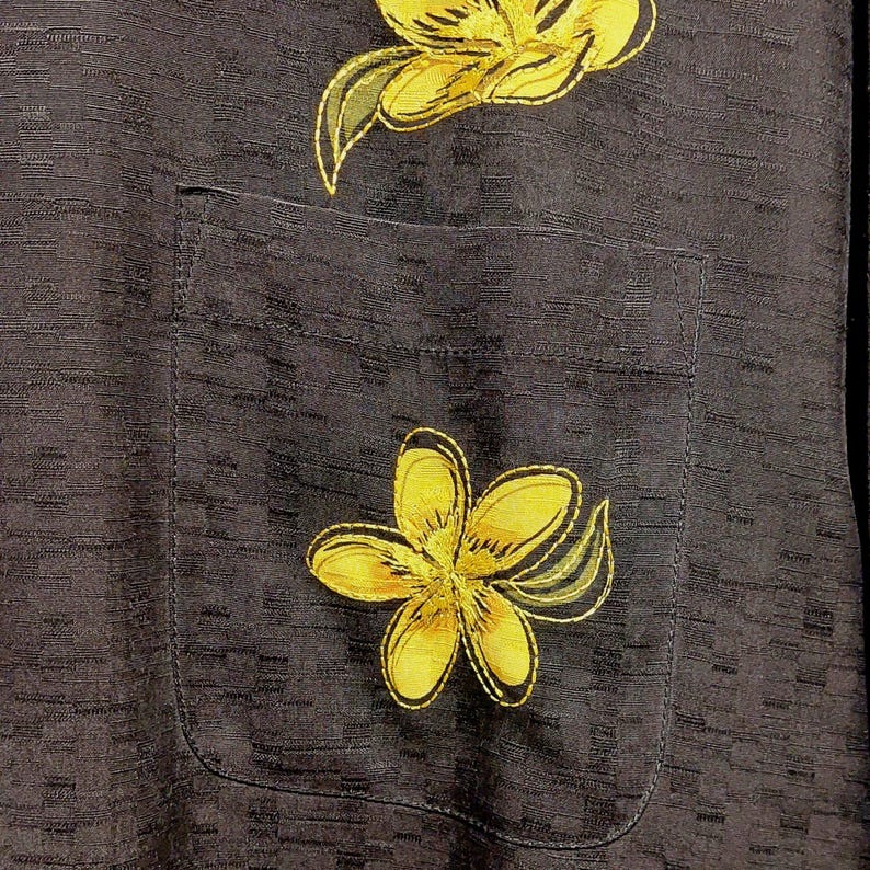 May include: Close-up of a black shirt with a pocket, featuring embroidered yellow flowers with green accents. The shirt has a textured pattern and a classic pocket design, showcasing a tropical floral theme.