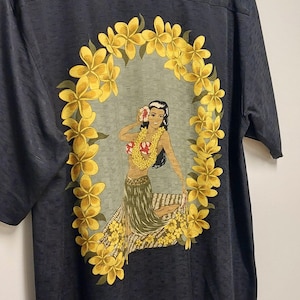 May include: Dark gray short-sleeved shirt featuring a Hawaiian-themed design. The graphic depicts a woman wearing a lei and a floral headdress, surrounded by a yellow flower wreath. The shirt has a textured appearance.