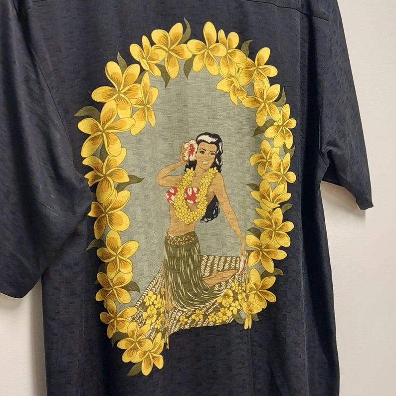 May include: Dark gray short-sleeved shirt featuring a Hawaiian-themed design. The graphic depicts a woman wearing a lei and a floral headdress, surrounded by a yellow flower wreath. The shirt has a textured appearance.