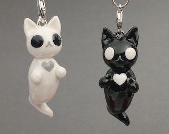 May include: Two cat charms with silver-toned lobster clasps. One charm is white with large black eyes and a grey heart on its chest. The other charm is black with large white eyes and a white heart on its chest. Both have smooth, glossy finishes.