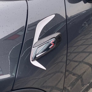 Könnte beinhalten: Nahaufnahme einer schwarzen Autotür mit einem weißen und roten "S" Emblem. Der Türgriff ist schwarz und verchromt mit einem weißen Akzent.