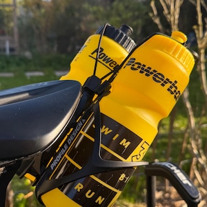 Könnte beinhalten: Zwei gelbe Wasserflaschen mit schwarzem Text, befestigt in einem schwarzen Flaschenhalter an einem Fahrrad. Auf den Flaschen steht "Powerbar" und "WIM". Der Halter hat die Aufschrift "Profile Design" und "Run".