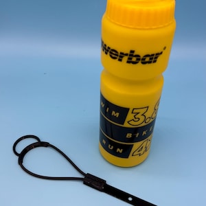 Op de afbeelding: Gele waterfles met zwarte accenten en het woord "werbar" erop gedrukt. De fles heeft een zwarte riem en een zwarte draagriem. De fles heeft de woorden "Swim, Bike, Run" en cijfers erop gedrukt.