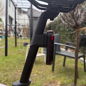 Könnte beinhalten: Eine schwarze Fahrradsattelstütze mit einem roten und schwarzen Rücklicht. Der Sattel ist schwarz und der Reifen ist unten sichtbar. Der Hintergrund zeigt einen Garten mit einem Haus und Bäumen.