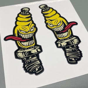 Könnte beinhalten: Zwei gelbe und schwarze Zündkerzen-Aufkleber mit einem Comic-Gesicht und einer roten Zunge. Die Zündkerzen haben einen schwarzen Umriss.