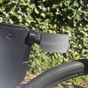 Op de afbeelding: Close-up van een zwart fietsonderdeel met een rechthoekig, getextureerd oppervlak. Het onderdeel is bevestigd aan een wit en zwart frame, met een deel van een zwarte band zichtbaar. Groene bladeren vormen de achtergrond.