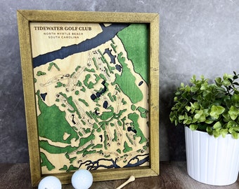 May include: A wooden framed map of Tidewater Golf Club in North Myrtle Beach, South Carolina. The map is laser cut and shows the layout of the golf course. The frame is a dark brown wood with a gold tint. There are golf balls and tees in the foreground.