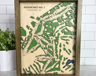 May include: A wooden framed wall art piece featuring a 3D laser cut map of the Pinehurst No. 2 golf course in Pinehurst, North Carolina. The map is green and brown and shows the layout of the golf course with numbered holes.