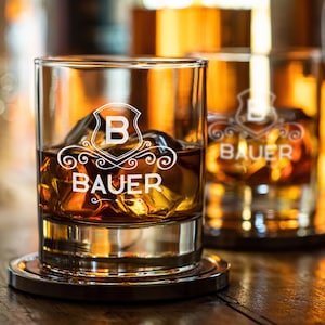 May include: Two clear glass tumblers with a personalized design. The glasses are filled with amber liquid and ice cubes. The design features a white shield with the letter "B" and the word "BAUER" underneath.