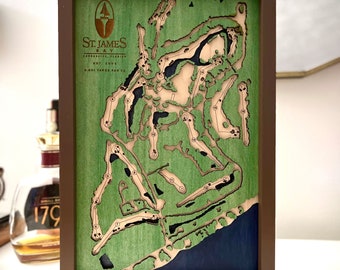 May include: A wooden golf course map of St. James Bay in Carrabelle, Florida. The map is framed in a brown wood frame and features a green background with a blue water feature. The course is outlined in white and numbered 1 through 18.