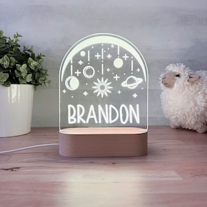 May include: A white acrylic night light with a crescent moon, stars, planets, and a sun design. The name "BRANDON" is written below the design. The night light is on a wooden base.