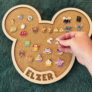 May include: A Mickey Mouse-shaped corkboard with a wooden frame, displaying various enamel pins. The board has the name "ELZER" engraved at the bottom. The pins feature cartoon characters and designs. The board is on a green surface.