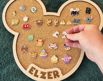 Puede incluir: Un corcho con forma de Mickey Mouse con un marco de madera, que muestra varios pines de esmalte. El tablero tiene el nombre "ELZER" grabado en la parte inferior. Los pines presentan personajes y diseños de dibujos animados. El tablero está sobre una superficie verde.