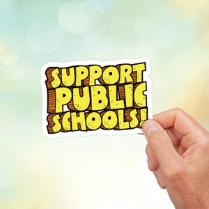 Puede incluir: Una pegatina blanca con las palabras "SUPPORT PUBLIC SCHOOLS!" en letras amarillas y naranjas. Las letras están en una fuente de estilo de dibujos animados.