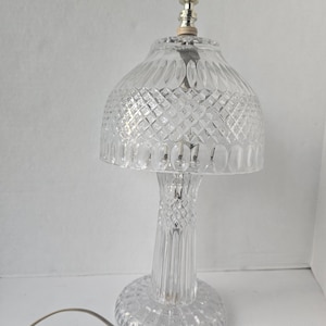 May include: A clear glass table lamp with a diamond pattern on the shade and base. The lamp has a cylindrical stem and a round base. The top of the lamp has a gold-colored finial. The lamp is plugged into an electrical outlet.