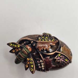 May include: A detailed, bronze-toned brooch featuring a bee and foliage design. The bee has green and orange accents, while the leaves and berries are adorned with colorful rhinestones. The brooch is oval-shaped and has a vintage aesthetic.