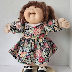 May include: A vintage doll with brown curly hair and a smiling face. The doll wears a floral print dress with a white lace collar and black shoes with white bows. The dress features a black background with pink, red, yellow, and blue flowers.