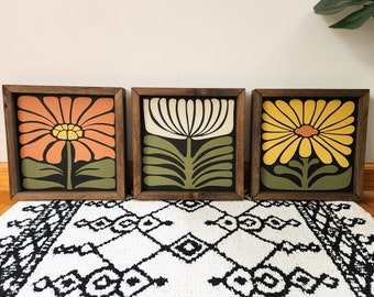 May include: Three square wooden framed wall art pieces featuring graphic floral illustrations. The left panel shows an orange flower, the center panel a cream flower, and the right panel a yellow flower, all set against dark backgrounds with green stems.