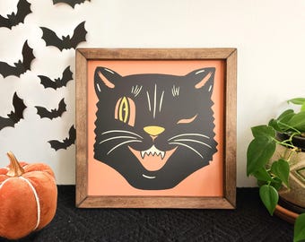 May include: Square framed Halloween art featuring a winking black cat with a yellow eye and a toothy grin, set against a peach background. The wooden frame adds a rustic touch. Decorative bats and a pumpkin are also visible.