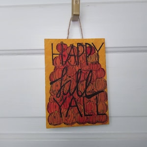 Puede incluir: Un letrero de madera amarillo con el texto "Happy Fall Y'all" escrito en negro. El letrero está decorado con una pila de calabazas naranjas y rojas.