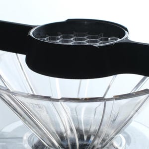 Könnte beinhalten: Ein klarer, kegelförmiger Kaffeezubereiter mit schwarzem Griff und Sockel. Der Zubereiter hat eine geriffelte Innenseite und sitzt auf einer klaren, kreisförmigen Basis. Der schwarze Griff hat ein wabenartiges Design.