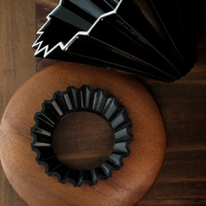 Puede incluir: Un abanico de papel origami negro y un anillo de origami negro se muestran sobre una superficie de madera. El abanico está parcialmente abierto, revelando su diseño plisado y el texto "ORIGAMI JAPAN". El anillo tiene un borde festoneado.