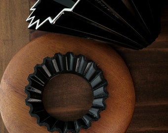 Könnte beinhalten: Ein schwarzer Origami-Papierfächer und ein schwarzer Origami-Ring werden auf einer Holzoberfläche gezeigt. Der Fächer ist teilweise geöffnet und zeigt sein gefaltetes Design und den Text "ORIGAMI JAPAN". Der Ring hat einen gewellten Rand.