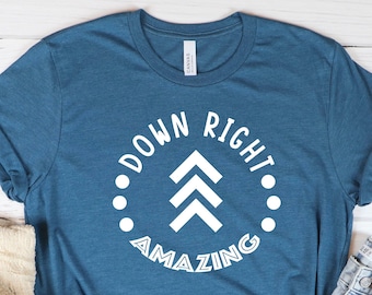 May include: Teal t-shirt with the text "DOWN RIGHT AMAZING" in a white circular design. The shirt is paired with denim shorts, white trainers with red trim, and a beige jumper. A mug with heart designs and a small plant are also in the image.