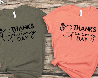 Może przedstawiać: Dwie koszulki z napisem "Thanks Giving Day" i małym obrazkiem rogu obfitości. Koszulka po lewej stronie jest oliwkowo zielona, a koszulka po prawej stronie jest koralowa.