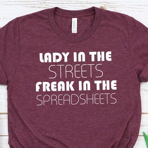 May include: Burgundy t-shirt with white text that reads "Lady in the Streets Freak in the Spreadsheets". The shirt is lying on a white wooden surface with a pair of white trainers and a bouquet of pink flowers.
