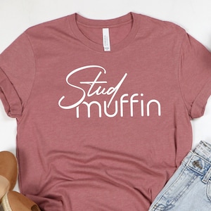 May include: A mauve t-shirt with the text "Stud muffin" in white lettering. The shirt is laid flat on a white surface with a pair of brown sandals and denim shorts.