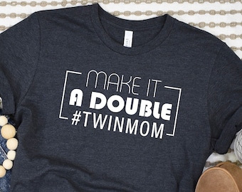 Könnte beinhalten: Ein dunkelgraues T-Shirt mit weißem Text, der "Make it a double #twinmom" lautet. Das Shirt ist auf einer hellbraunen und weißen gestreiften Oberfläche mit blauen Jeans, einer Holzperlenkette und braunen Wildlederschuhen abgebildet.