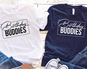 Könnte beinhalten: Zwei T-Shirts, eines weiß und eines marineblau, mit dem Text "Birthday Buddies" in einem schwarzen Kasten. Die Shirts liegen auf einer Holzoberfläche mit Jeans und Schuhen.