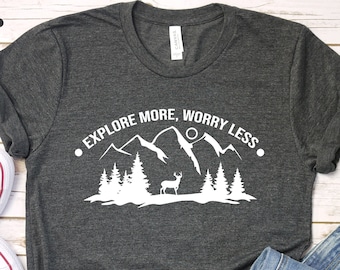 May include: A dark grey t-shirt with a white graphic design of mountains, trees, and a deer. The text "Explore More, Worry Less" is printed in white on the shirt.