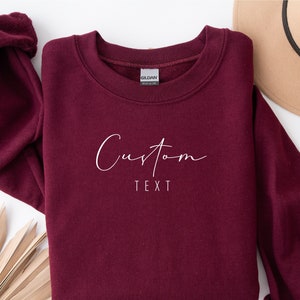 Może przedstawiać: Bordowa bluza z białym napisem „Custom Text” w ozdobnym, pochylonym stylu.