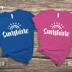 May include: Two t-shirts with a white sun and the word "Sunshine" printed on them. The shirt on the left is royal blue and the shirt on the right is hot pink.