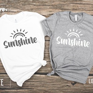 May include: Two t-shirts with the word "Sunshine" and a sun graphic. One t-shirt is white with a gray graphic and the other is light gray with a white graphic.