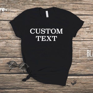 May include: Black t-shirt with the text "CUSTOM TEXT" printed in white on the front. The shirt is laid flat on a wooden surface.
