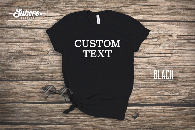 May include: Black t-shirt with the text "CUSTOM TEXT" printed in white on the front. The shirt is laid flat on a wooden surface.