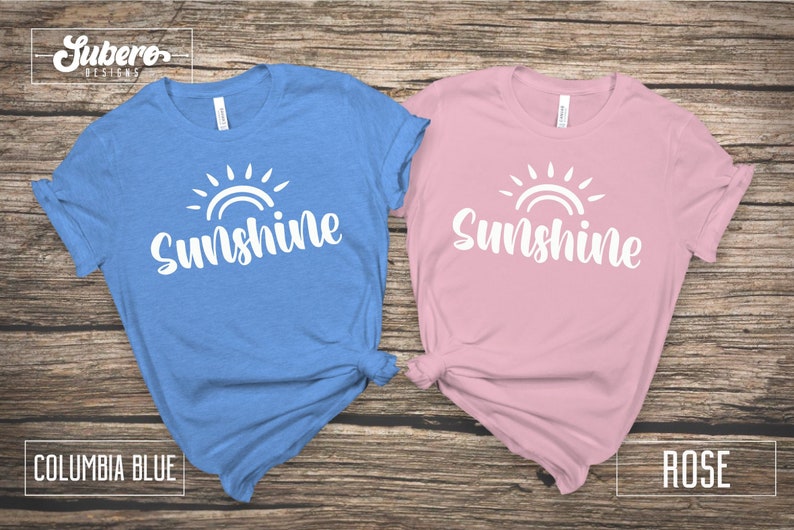 May include: Two t-shirts with the word "Sunshine" and a stylized sun graphic. The shirt on the left is Columbia blue and the shirt on the right is rose.