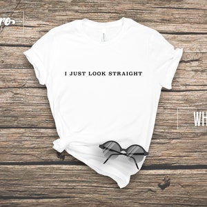 May include: White t-shirt with the text "I JUST LOOK STRAIGHT" printed on the front. The shirt is laid flat on a wooden surface with a pair of sunglasses on top of it.