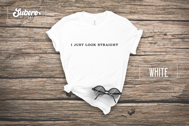 May include: White t-shirt with the text "I JUST LOOK STRAIGHT" printed on the front. The shirt is laid flat on a wooden surface with a pair of sunglasses on top of it.