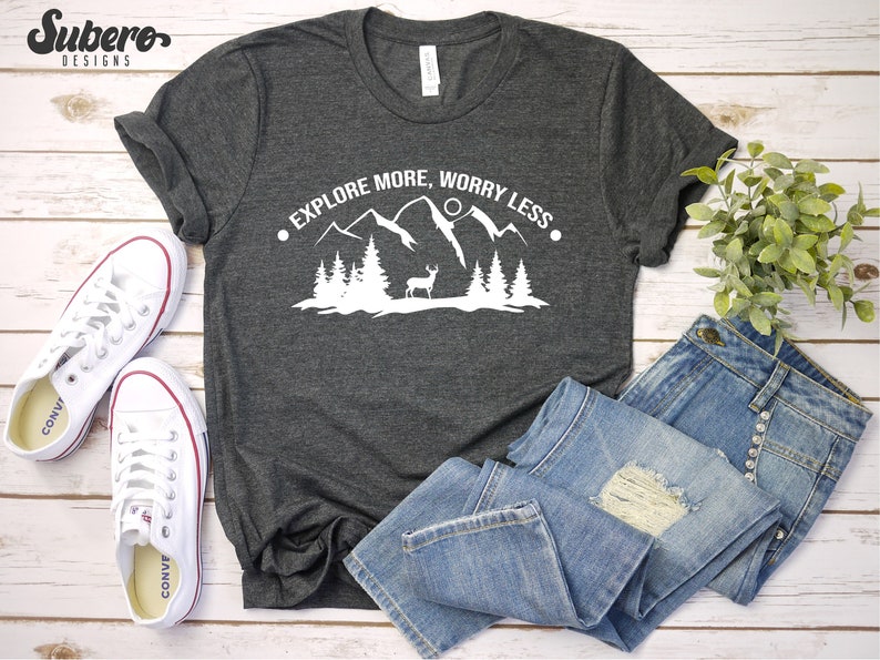 K&ouml;nnte beinhalten: Ein dunkelgraues T-Shirt mit einem wei&szlig;en Grafikdesign von Bergen, B&auml;umen und einem Reh. Der Text "Explore More, Worry Less" ist in Wei&szlig; auf das Shirt gedruckt.