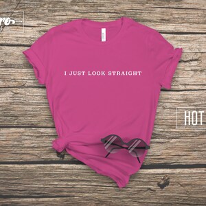 May include: Hot pink t-shirt with the text "I just look straight" printed in white on the front. The shirt is laying flat on a wooden surface with a pair of sunglasses on top of it.