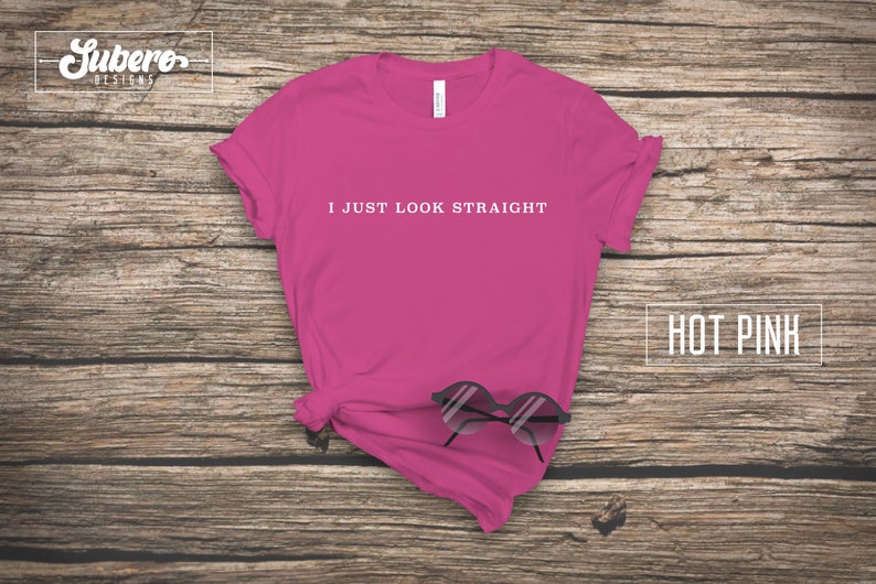 May include: Hot pink t-shirt with the text "I just look straight" printed in white on the front. The shirt is laying flat on a wooden surface with a pair of sunglasses on top of it.